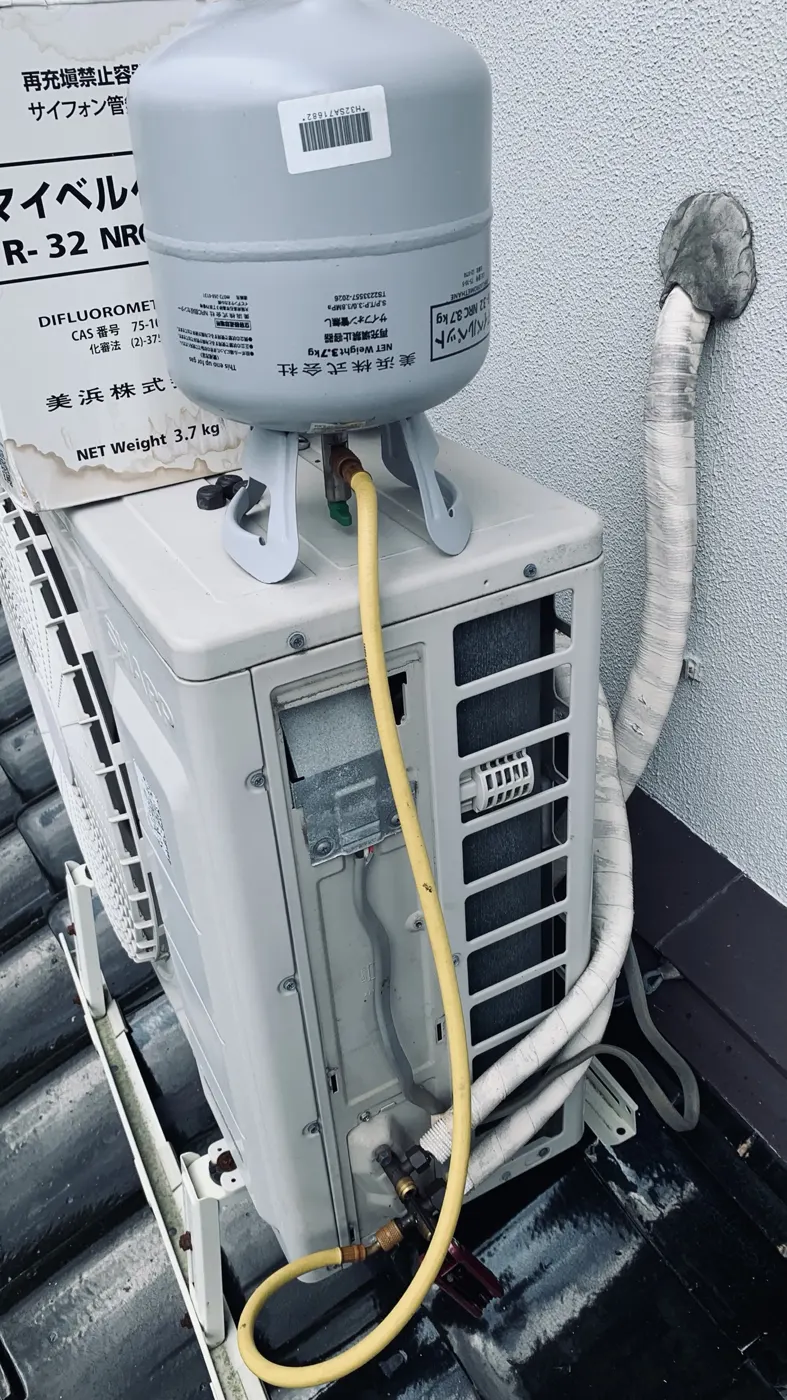 屋根の上の室外機に冷媒ガス(R-32)のボンベを載せ、黄色いホースでつないで補充している